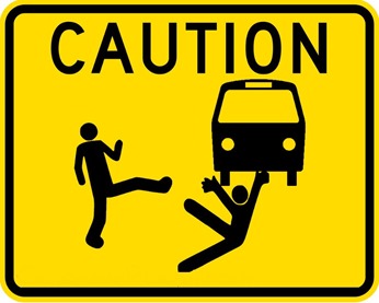 cartello con la scritta CAUTION e figure stillizzate di una persona che con un calcio ne fa cadere un’altra davanti a un autobus che sopraggiunge
