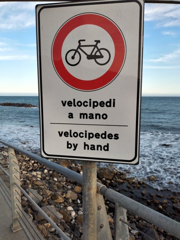 Cartello con segnale di divieto di transito a biciclette e scritte VELOCIPEDI A MANO e in pseudoinglese VELOCIPEDES BY HAND